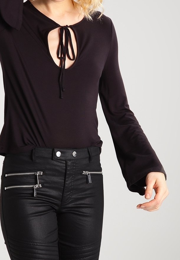 Kvinna som bär en svart långärmad topp med en nyckelhålsklippning och svarta byxor med flera silverdragkedjor och knappar.