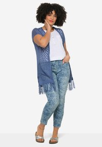 Vaaleansininen neulecardigan, jossa on avonainen etuosa, geometrisia kuvioita ja fringe-tyylinen helma, päällä valkoinen toppi ja kuvioidut siniset farkut.