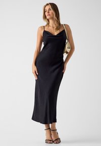 Μαύρο σατέν φόρεμα με λεπτές τιράντες και ντεκολτέ καρφωτό. Το μοντέλο φοράει χρυσή τσάντα και μαύρα ψηλοτάκουνα σανδάλια, με ένα ουδέτερο φόντο.