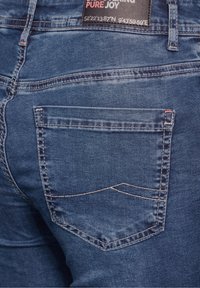 Mörkblå denimjeans med en bakficka med kontrasterande sömmar och en etikett. Texturen ser något sliten och mjuk ut.