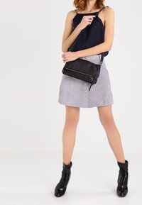 Borsa a tracolla nera, gonna in suede grigia con bottoni frontali, top senza maniche blu scuro e stivaletti neri, tutti su uno sfondo neutro.