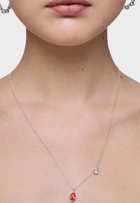 Collar de cadena de plata con un colgante de piedra preciosa roja y un pequeño acento plateado, llevado en un cuello descubierto con tono y textura de piel visibles.