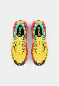 Zapatillas deportivas amarillas brillantes con detalles en negro y suela roja vibrante. Parte superior de malla con patrones intrincados y cordones turquesa. Branding de HOKA visible.