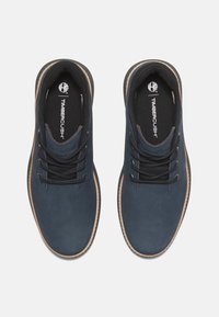 Zapatos de cuero azul oscuro con cordones negros, punta redonda y suelas de goma beige. Presenta un cuello acolchado y un interior texturizado. Logotipo de Timberland visible.
