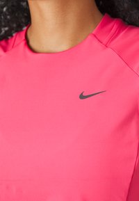 Sportovní top Nike v jasně růžové barvě, s kulatým výstřihem, krátkými rukávy a černým „swoosh“ logem na hrudi. Hladká, lehká tkanina.