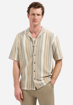 Mann trägt kurzärmliges Hemd mit beige-grau-weißer Zickzack-Muster, steht mit einer Hand in der Tasche vor weißem Hintergrund.