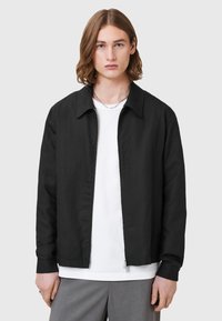 Svart jacka med krage, framdragkedja, sidofickor och en slät yta, bärs över en vit skjorta. Avslappnad design.