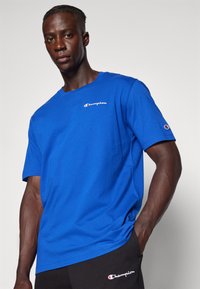 T-shirt en coton bleu à manches courtes, avec un logo "Champion" blanc sur la poitrine et une étiquette logo sur la manche.