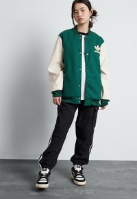 Green and cream varsity jacket with snap buttons, black cargo pants with white stripes, and black and white sneakers.