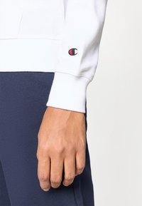 Λευκό φούτερ με ριμπ μανσέτα και μικρό κόκκινο και μαύρο λογό τυπο στο αριστερό μανίκι, φορεμένο πάνω από μπλε σκούρο παντελόνι. Υφασμάτινη υφή, casual εφαρμογή.