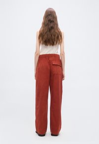 Femme aux cheveux longs et ondulés debout, portant un pantalon ample orange brûlé avec une poche arrière et un haut sans manches clair sur un fond uni.