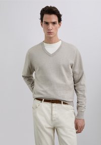 Beiger V-Ausschnitt-Pullover aus weichem Material, mit langen Ärmeln und gerippten Bündchen, kombiniert mit hellen Hosen und einem geflochtenen braunen Gürtel.