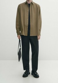 Braune Button-Up-Hemd, darunter ein schwarzes Rundhalsoberteil, schwarze Hose, schwarze Schuhe, hält eine kleine schwarze Tasche, steht vor einem neutralen Hintergrund.