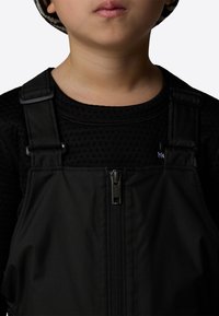 Black overalls with adjustable straps and a front zipper, paired with a textured black shirt. Features a small white logo on the shirt.