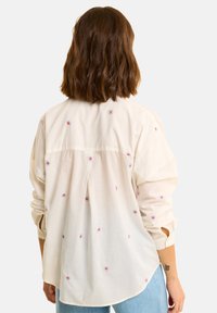 Camicia beige a maniche lunghe con ricami floreali colorati, vestibilità rilassata, polsini con bottoni e orlo curvo. Tessuto leggero e morbido.