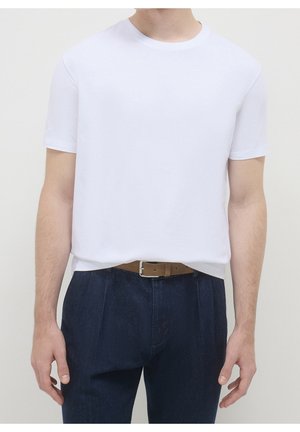 Hombre con camiseta blanca lisa de manga corta metida dentro de pantalones azul oscuro con cinturón marrón, de pie frente a un fondo neutro.