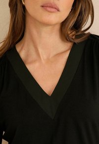 Schwarzes Oberteil mit tiefem V-Ausschnitt und grünen Akzenten. Weicher Stoff mit leichter Struktur. Ärmel an den Schultern gerafft für zusätzliche Details.