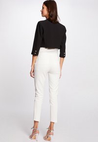 Schwarze kurz geschnittene Jacke mit Dreiviertelärmeln und goldenen Knopfakzenten, kombiniert mit weißen, figurbetonten Hosen und silbernen High-Heels.