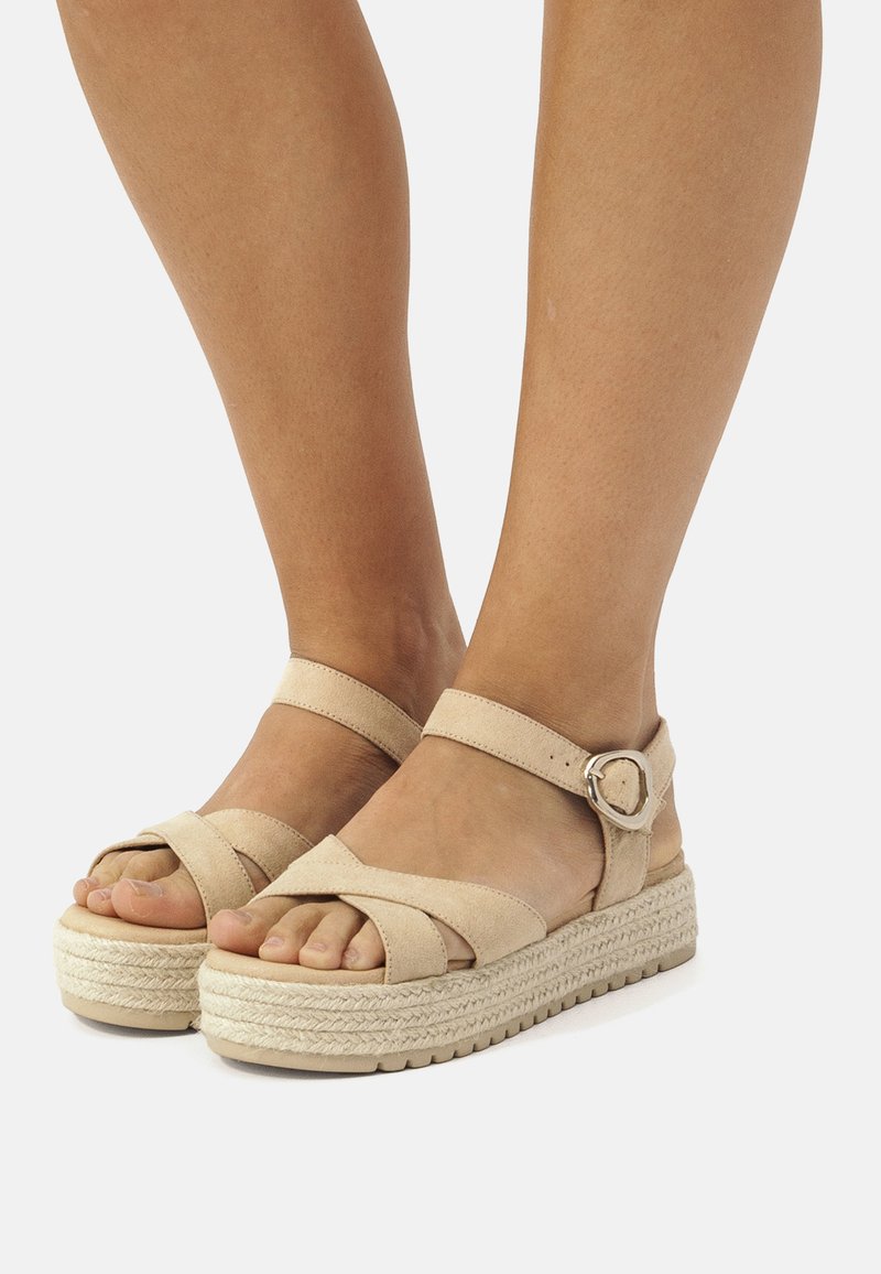 Beige espadrille-sandaalit, joissa on ristikkäiset mokkapohja-nauhat, nilkkamikä ja kudottu juuttipohja. Avoin varvasmuotoilu esittelee varpaita.
