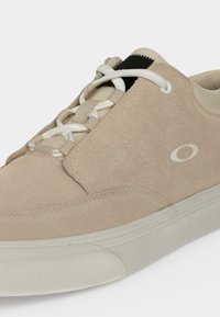 Baskets en daim beige avec lacets blancs, semelle blanche texturée et petit logo ovale sur le côté.