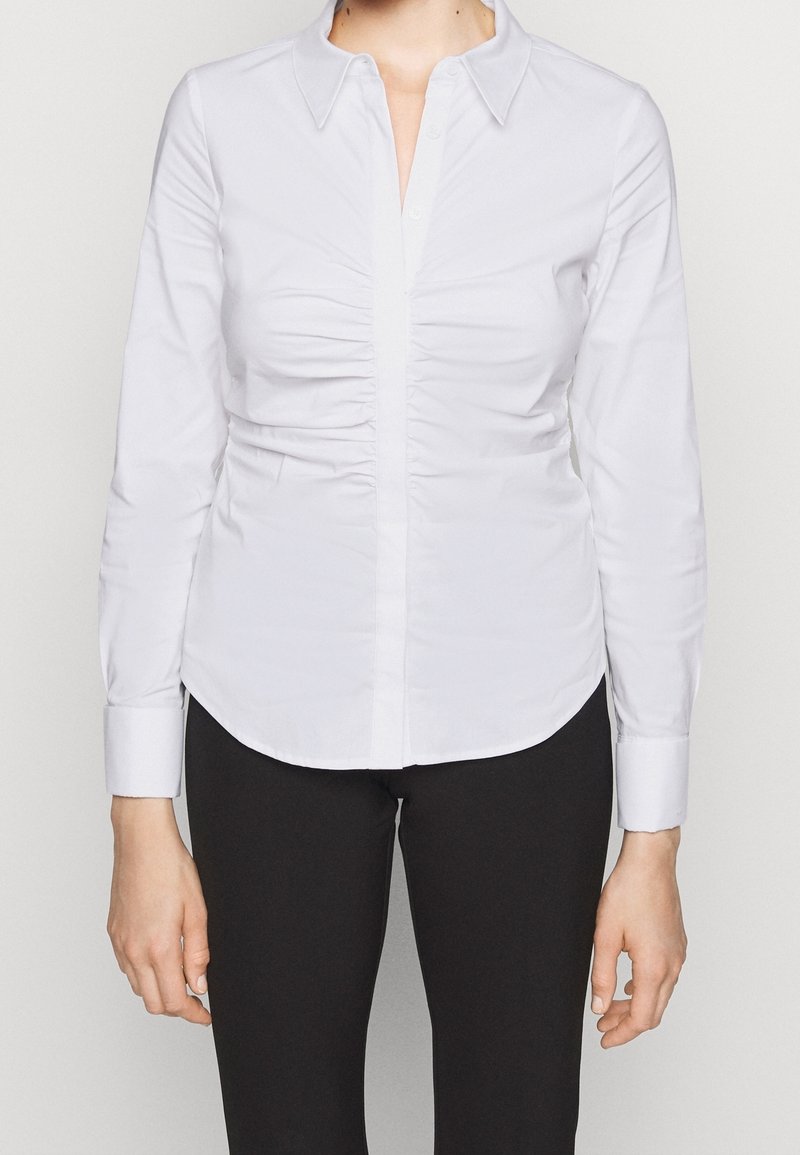 Chemise blanche à manches longues froncée avec boutons, portée avec un pantalon noir, montrée du cou aux hanches sur un fond uni.