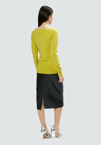 Un maglione aderente a maniche lunghe di un brillante colore giallo abbinato a una gonna a matita in pelle nera con uno spacco posteriore, indossato con tacchi alti argentati.
