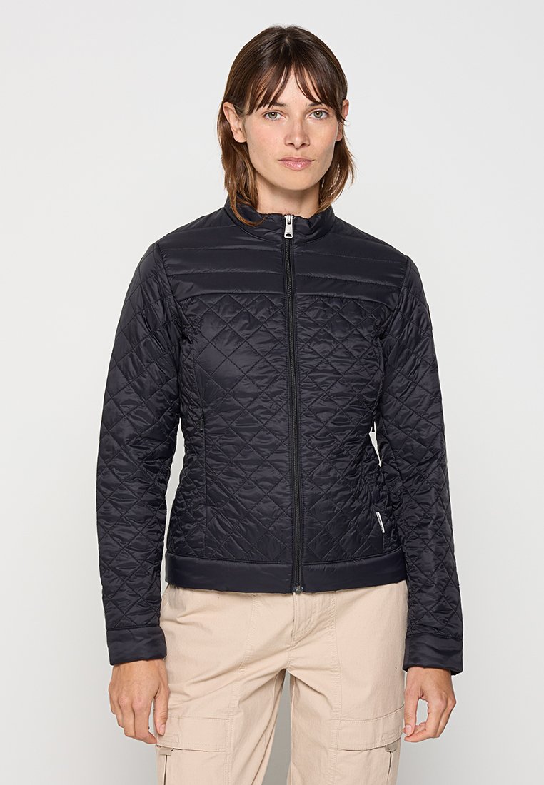 Frau in einer schwarzen gesteppte Jacke mit Reißverschluss und beigen Cargo-Hosen, die vor einem schlichten hellen Hintergrund steht.