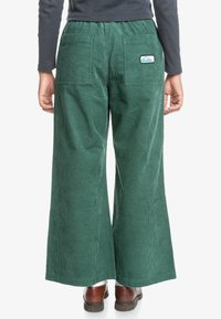 Wide-leg corduroy pants in deep green, featuring a stretch waistband, back pockets, and a small logo patch. Smooth, textured fabric.