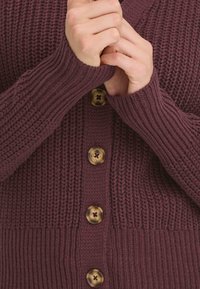 Burgundy neulejumper, jossa on ribattu pinta, V-pääntie, pitkät hihat ja kuusi isoa kultasävyistä nappia edessä.