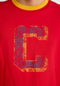 Sarkans kokvilnas T-krekls ar dzeltenu ribotu apkakli. Priekšpusē ir liels, nolietots zils un zelta burts "C" dizains.