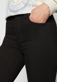 Personne portant un pantalon noir, la main dans la poche avant droite et un sweat crème avec un emblème bleu du Kansas.