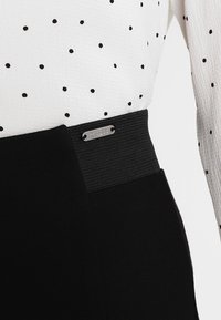 Zwarte rok met een brede elastische tailleband en een zilverkleurig logolabel. Het bovenstuk is een witte, geribbelde, lange mouwen blouse met zwarte stippen.