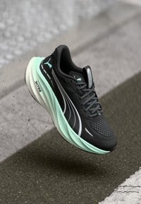 Chaussure de course Puma noire et vert menthe suspendue au-dessus de l'asphalte avec un amorti Nitro visible sur la semelle.