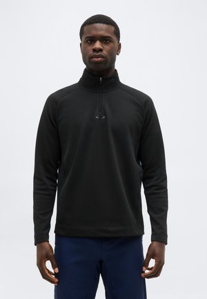 Mann trägt einen schwarzen Pullover mit Viertel-Reißverschluss und dunkelblaue Hose, steht mit entspannten Armen vor einem einfarbigen hellen Hintergrund.