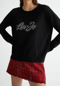 Maglione nero con scollatura rotonda, caratterizzato da un logo argentato luccicante. Abbinato a una gonna a quadri rossa con una texture e bordi frangiati.