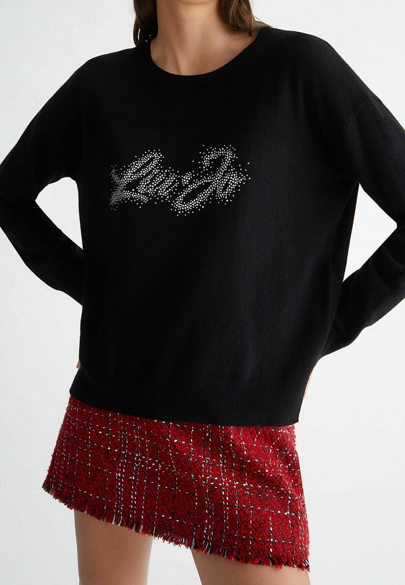 Maglione nero con scollatura rotonda, caratterizzato da un logo argentato luccicante. Abbinato a una gonna a quadri rossa con una texture e bordi frangiati.