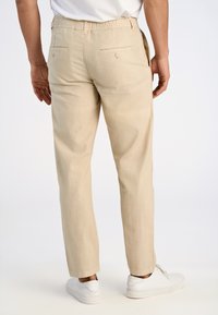 Beige linnen broek met een elastische tailleband, twee achterzakken en een taps toelopend ontwerp. Gekleed met witte sneakers.