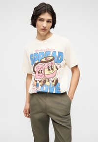 Νεαρός φοράει κρεμ μπλουζάκι με χαρακτήρες καρτούν τοστ και βαζάκι και το κείμενο «Strawberry Goodness SPREAD the LOVE.»