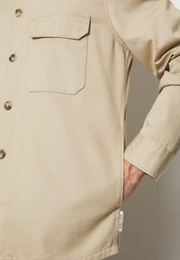 Beige shirt jacket made from cotton, featuring a patch pocket, button closure, and side slits. Label visible at the hem.