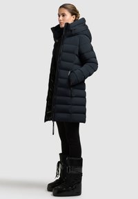 Mujer de pie con abrigo de plumas negro con capucha, leggins negros y botas de invierno negras, contra un fondo claro y liso.