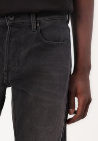 Jeans de mezclilla gris oscuro con corte recto, que presentan un diseño clásico de cinco bolsillos y detalles de costura. Cierre de botón y cremallera en la cintura.