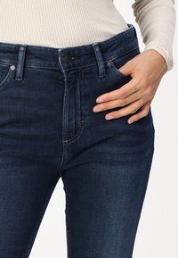 Los pantalones vaqueros de mezclilla azul oscuro presentan un diseño ajustado con una cintura de altura media, bolsillos frontales y una sutil textura desgastada a lo largo de los muslos.