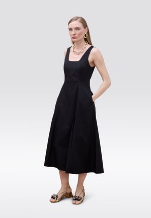 Vestido midi de algodón negro con escote cuadrado, cuerpo ajustado, falda acampanada y bolsillos laterales; combinado con sandalias doradas con adornos.