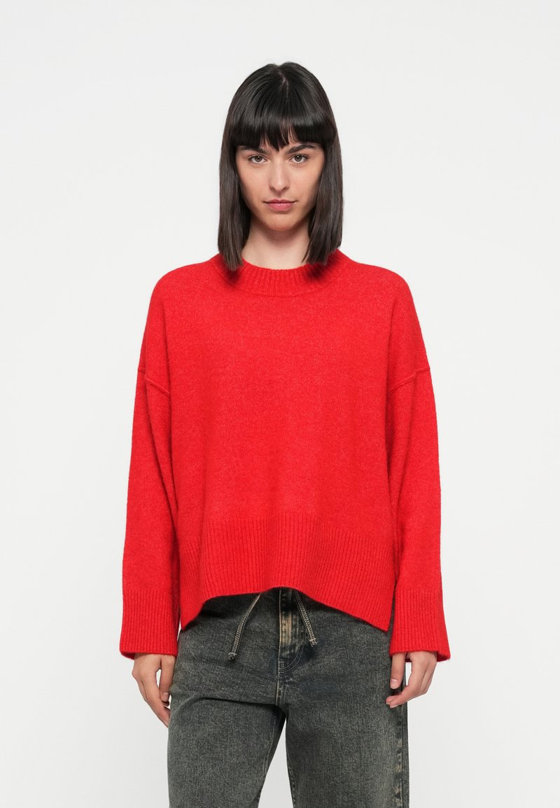 Rode gebreide trui met een geribbelde ronde halslijn en brede mouwen, gestyled met een relaxte pasvorm. Gedragen over donkere, gestructureerde jeans.