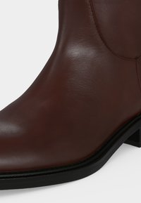 Gros plan d'une botte cheville en cuir marron avec un bout arrondi et un petit talon noir sur un fond clair uni.