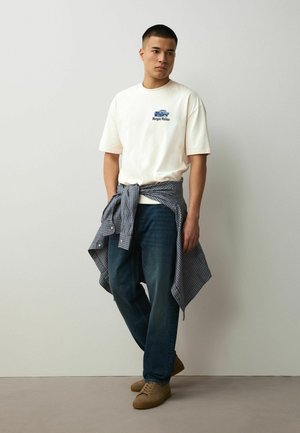 Giovane uomo in piedi contro un muro che indossa una t-shirt bianca con grafica, jeans scuri, scarpe color cuoio e una camicia azzurra a quadri legata in vita.