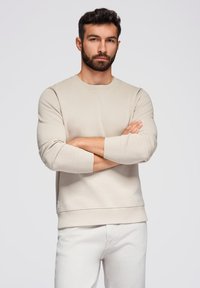 Felpa beige realizzata in morbido tessuto, con scollo rotondo e polsini a coste. Il modello incrocia le braccia, indossando pantaloni bianchi.