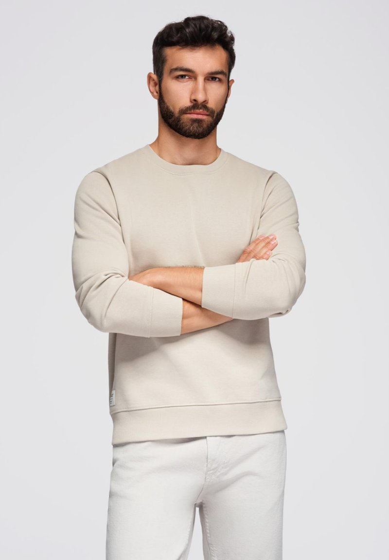 Felpa beige realizzata in morbido tessuto, con scollo rotondo e polsini a coste. Il modello incrocia le braccia, indossando pantaloni bianchi.