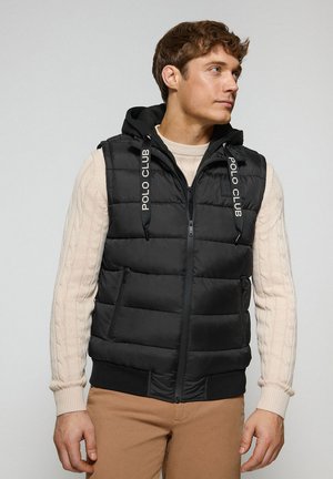 Black padded vest with a hood, featuring a zip front, two side pockets, and branded drawstrings. Worn over a light beige cable-knit sweater.