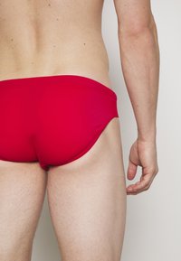 Rode zwemslips van rekbare stof, met een lage taille en een gladde textuur. Zichtbare stiksels langs de naden en de beenopeningen.